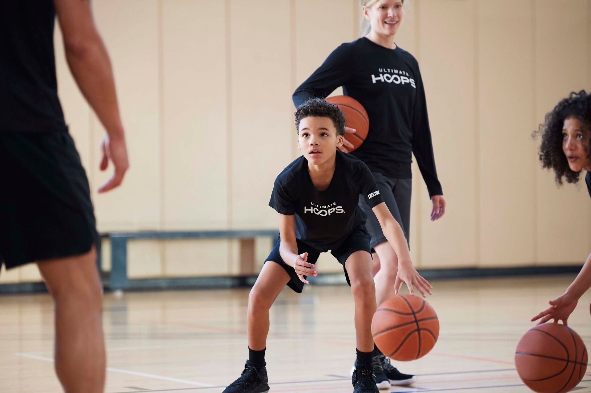 Children and adult playing basketball indoors, wearing black shirts with 'Ultimate Hoops' logo. Children and adult playing basketball indoors, wearing black shirts with 'Ultimate Hoops' logo.